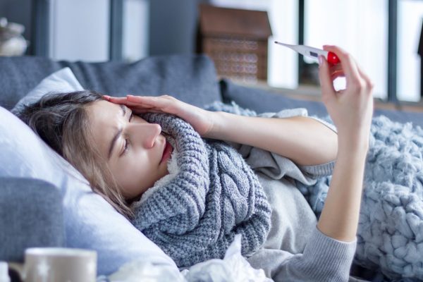 Sick exhausted girl in scarf is lying in bed wrapped in blanket. Young woman with fever and headache is measuring temperature with thermometer, treated at home. Winter cold and flu concept.