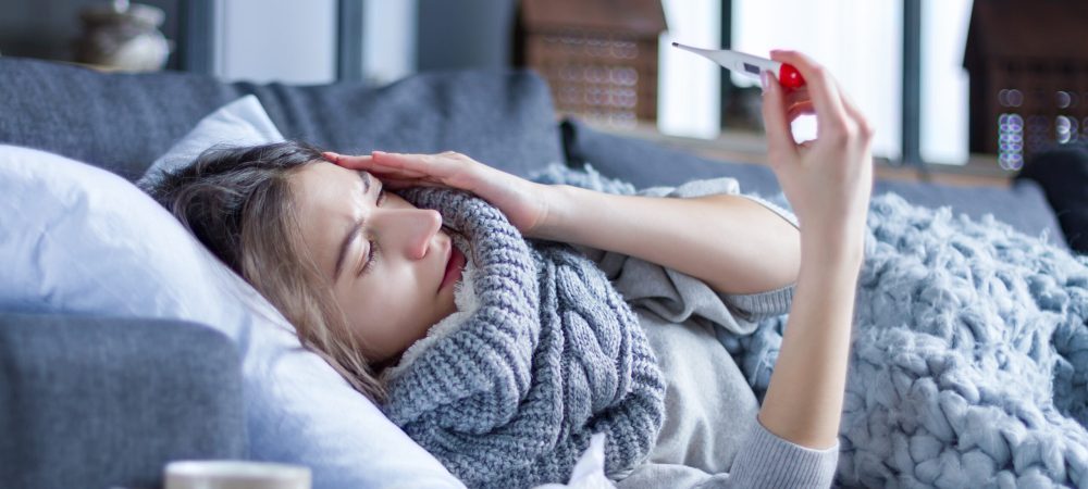 Sick exhausted girl in scarf is lying in bed wrapped in blanket. Young woman with fever and headache is measuring temperature with thermometer, treated at home. Winter cold and flu concept.